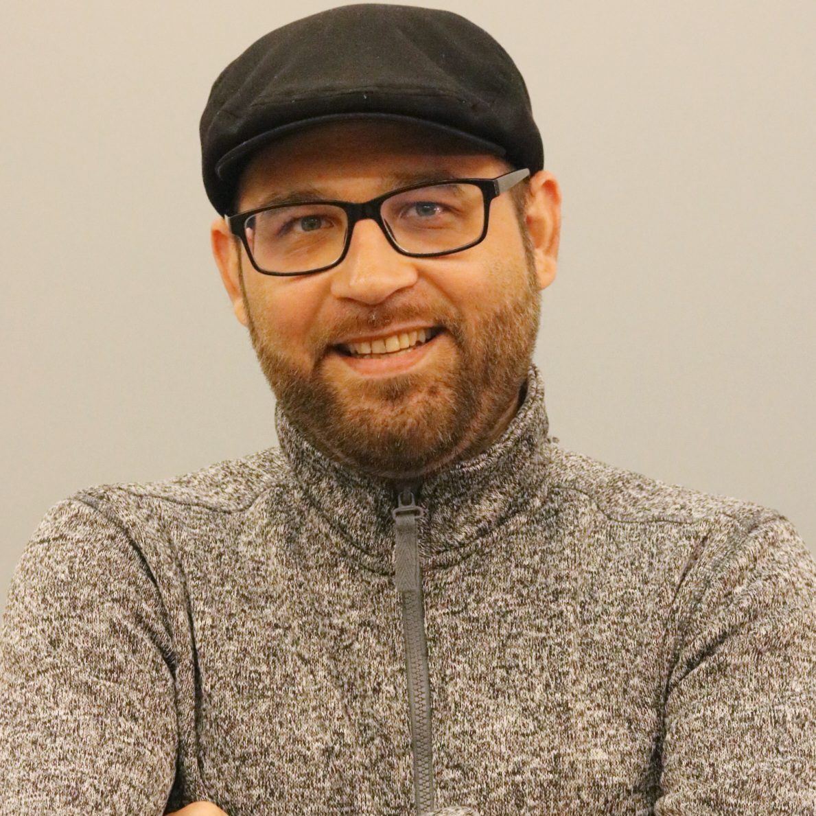 Memo Monge, instructor at Creating Coding Careers, smiling with arms crossed, wearing a gray zip-up sweater and a black cap, against a neutral background.